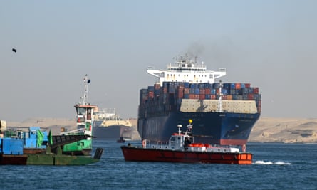 A cargo vessel leaves the Suez Canal to enter the Red Sea at Ismailia, Egypt: it is seen from behind, heavily laden with containers, as a smaller red pilot vessel escorts it; another large vessel and marine machinery are seen in the background