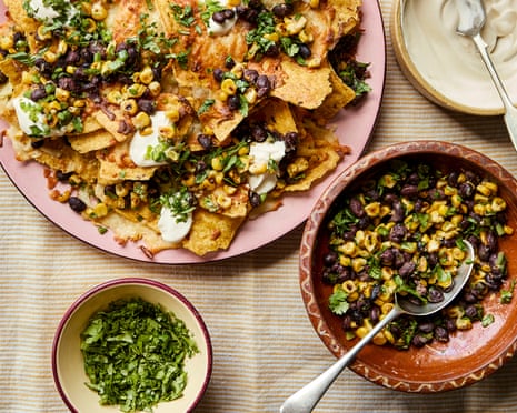 A pink plate of nachos topped with black beans, corn, herbs, and sour cream, surrounded by small bowls