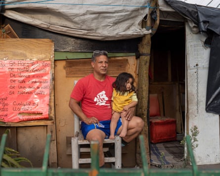 A man sits on a chair outside a makeshift camp with his young daughter on his lap