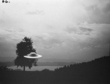 Black and white image of a saucer-shaped UFO above mountains with a tree behind