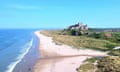 Bamburgh castle seen in the distance overlooking a long stretch of sandy beach and calm sea; the sky is blue and there is a small village seen behind the castle