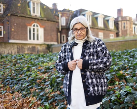 she is nearly 70 and has long white hair; she is standing in a garden and wears a heavy black and white cardigan over a white jumper