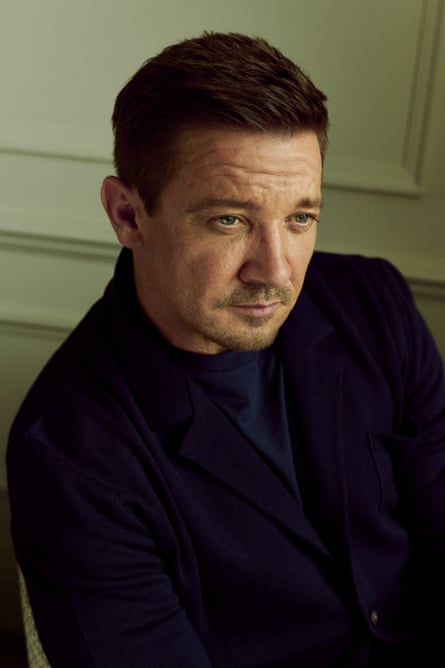 Portrait of Jeremy Renner sat in a chair wearing a dark t-shirt and jacket with a pale wood-panelled wall in the background