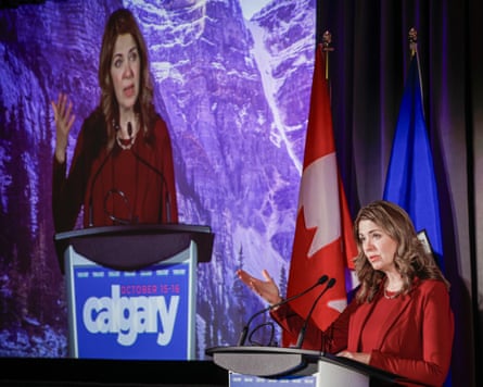 Alberta Premier Danielle Smith speaks at a podium beside flags, her image shown on a large screen