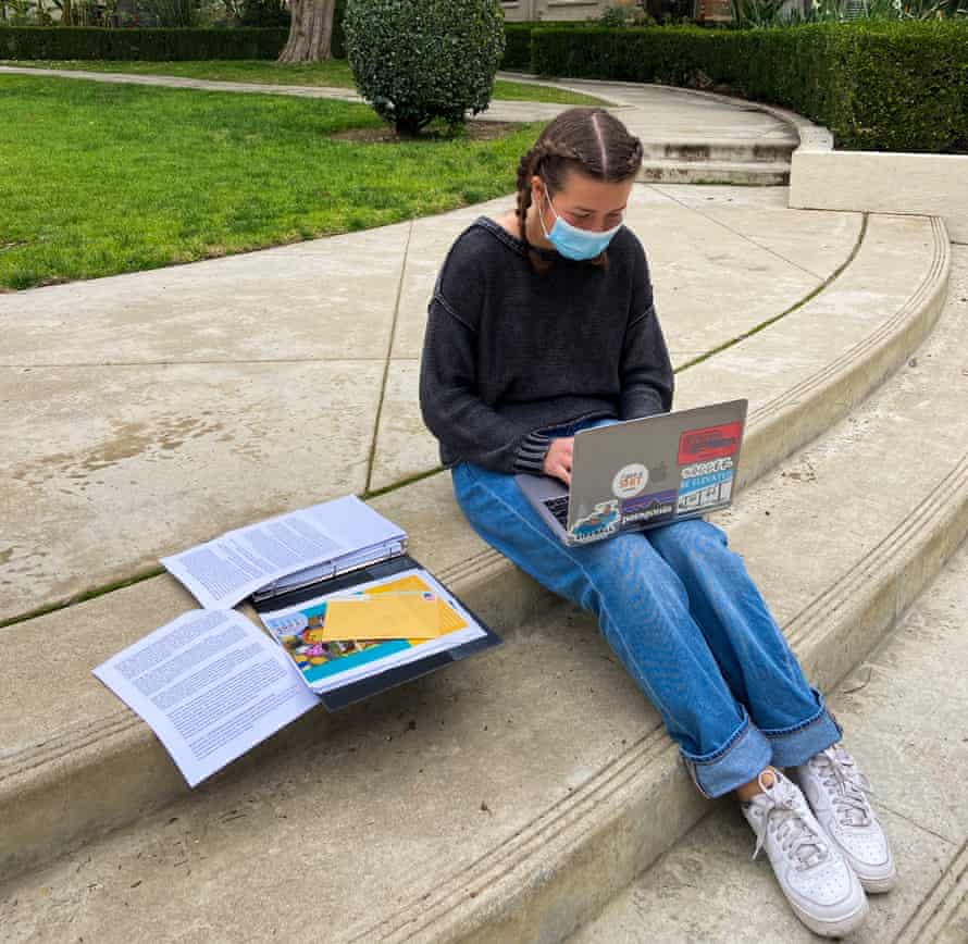 Maggie Callow, a young woman wearing jeans with her dark brown hair in two french braids, looks down at a laptop computer as she sits outside on the campus of Pomona College. A stack of papers are beside her.