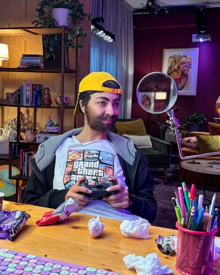 A woman dressed as a boy with beard and a cap on backwards sits at a table in a living room holding a games console