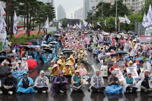a large public demonstration or protest taking place on a rainy day. Hundreds of people, many wearing colourful rain ponchos and holding umbrellas, are seated on a street in Seoul, South Korea.