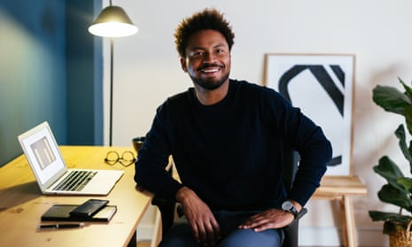 Positive black male in casual clothes smiling and looking at camera while sitting near table with laptop in workplace at home