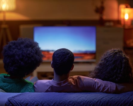 Back view of three people sitting on couch together and watching TV in dark room
