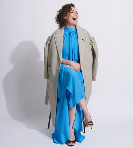 Olivia Colman in blue dressand beige coat, sitting on a chair, laughing, October 2021