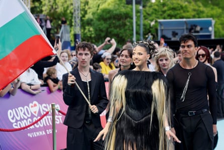 Dara walking in black and gold outfit with person holding Bulgarian flag and several other with spectators behind