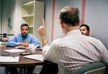 Three men sitting around an office desk, one with his back to the camera, holding a finger up