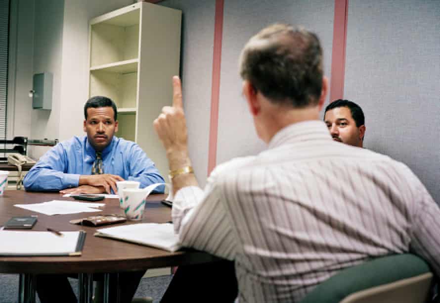 Three men sitting around an office desk, one with his back to the camera, holding a finger up