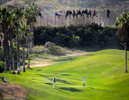 A golfer hits a tee shot as African migrants sit atop a border fence during an attempt to cross into Spanish territories between Morocco and Spain’s north African enclave of Melilla October 22, 2014