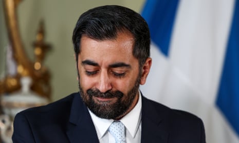 Humza Yousaf looking at the ground during a press conference. A Scottish flag is in the background.