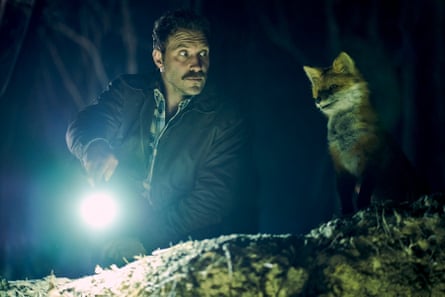 Outdoor shot at night featuring a young man with a moustache holding a flashlight, looking at an animatronic fox sitting beside him on a grassy hill.