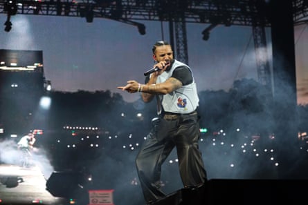 A bearded rapper singing on stage with black T-shirt and white vest and black trousers