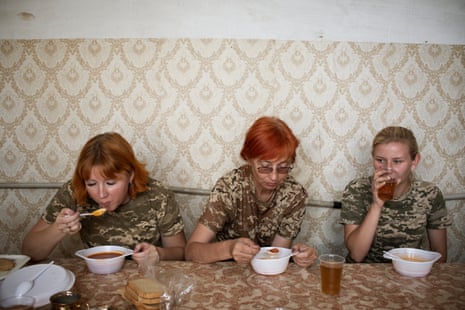 Three women wearing camouflage T-shirts sit in a row at a table eating food from white bowls