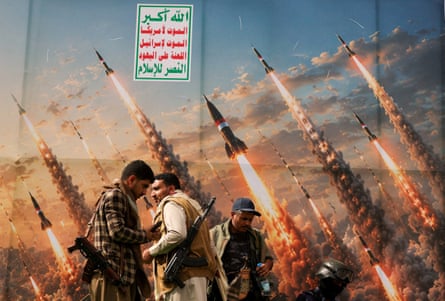 Men with rifles around their shoulders stand in front of a mural depicting missiles being fired into the sky