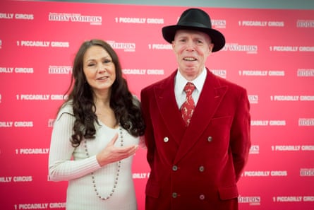 Angelina Whalley in a white dress and Gunther von Hagens in a red suit and black fedora pose for photos at an event