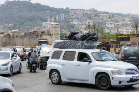 A traffic jam with cars and motorcycles queueing to leave the city. There are old stone walls and turrets alongside the road and the city spreads up a hillside in the background.