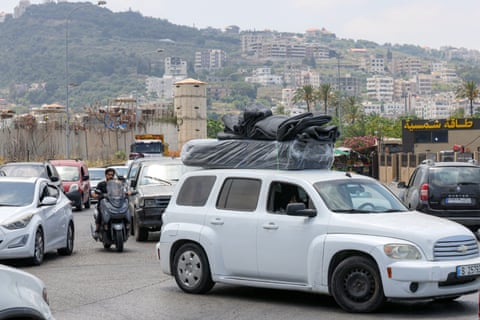 A traffic jam with cars and motorcycles queueing to leave the city. There are old stone walls and turrets alongside the road and the city spreads up a hillside in the background.
