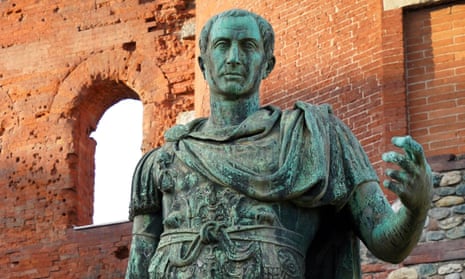 A closeup of an outdoor, oxidized bronze statue of a young Julius Caesar, wearing a cloak across his shoulders and holding up his left hand, as if at one point it held something. The statue stands before an old, very high brick wall with arch windows.