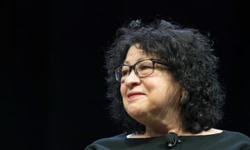 n older Latina woman with black hair and glasses, smiling and lit with a microphone attached to her blouse as if on a stage.