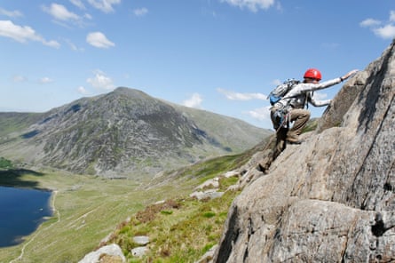 A climber in red helmet on a rocky ascent, with a mountain, green slopes and a lake in the background