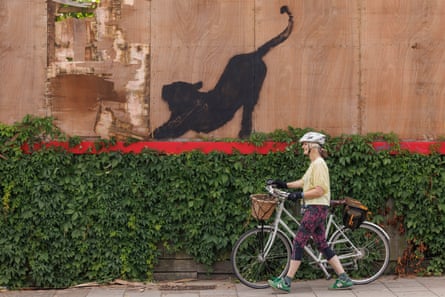 A silhouetted cat on a billboard, with a woman walking by pushing a bicycle