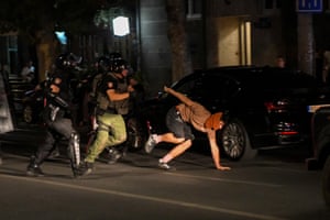 A man, his face covered by a reddish scarf, puts his hand down on the asphalt to try to stop himself falling to the ground as security forces in riot gear close in