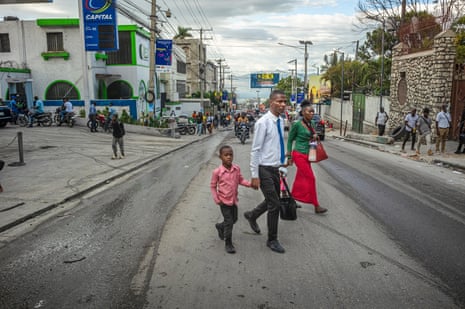 A man and a woman in smart dress cross a road, each holding the hand of a child