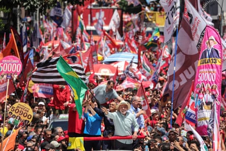 Lula waving Brazil flag from open-top bus among huge crowd