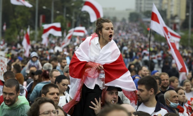 Protestor, draped in Belarusian flag, shouts from the shoulders of those in a large crowd.