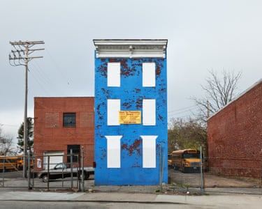 A tall blue house, windows overpainted, sits in a lot where school buses are parked on either side