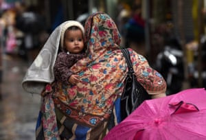A woman carries a young child during rainfall, an umbrella is in the foreground