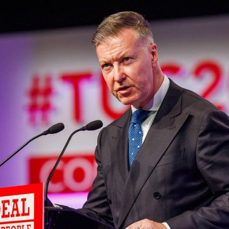 he wears a dark grey double-breasted jacket and blue tie with white spots and has short, combed-back greying hair.