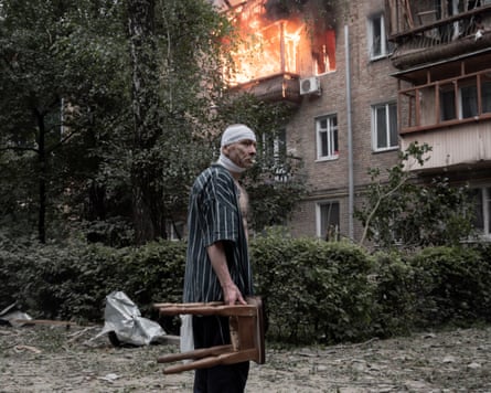 A man with a bandage on his head carrying a chair outside a burning apartment block,