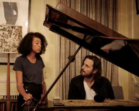 A man with long hair sits at a piano and watches a woman as she sings.