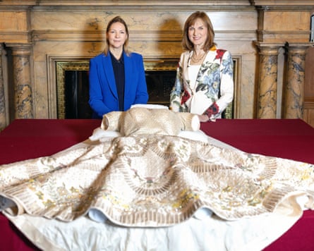 Fiona Bruce and the Royal Collections Curator, Caroline de Guitaut, stand infront of a red table, on which is laid out Queen Elizabeth II's coronation dress.