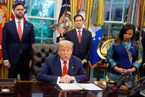 Trump seated in the Oval Office with two men standing either side of him, all wearing navy suits and red ties. An African woman stands beside Trump's desk