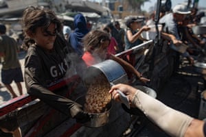 Palestinians, including children, hold pots as they queue to receive food from a kitchen, a girl in the foreground is having pasta poured into a bowl from a large saucepan