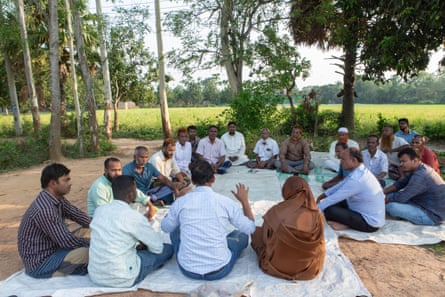 A group of men and women sit in a circle on rugs next to a field.