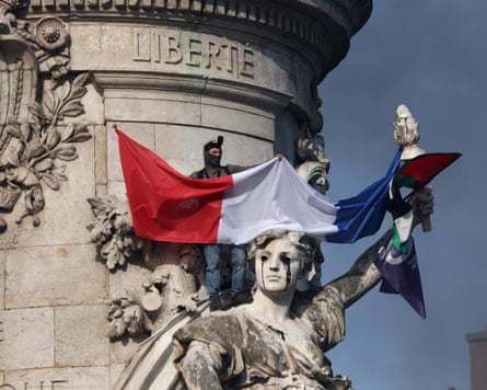 A masked protester stands on a statue holding a French flag beside a weeping stone figure