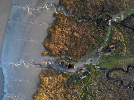 Aerial view of the Wyre estuary, where wrecked and decaying wooden boats sit on a green, reedy mudbank at a creek that forks and snakes inland.