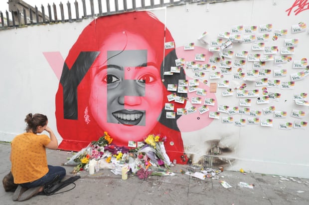 A woman kneels in front of wall with a mural of Savita Halappanavar, with notes on it and flowers left there 
