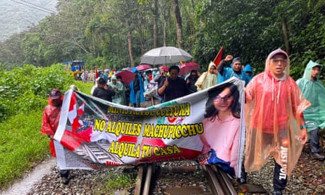 People in raincoats and rain ponchos hold umbrellas and a banner as they walk on rail tracks.