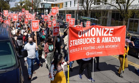 A crowd of people march down a city street holding red signs. The large banner being held by three people in front of the pack reads 'Unionize Starbucks & Amazon'.