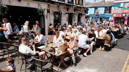 Drinkers sit at wooden tables in the street outside a cream-painted pub. It is hot and sunny and they are dressed in shorts, sunglasses and summer clothes.