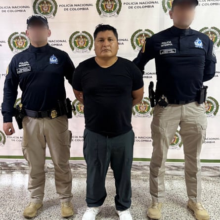 A stocky man in black clothing stands with his hands cuffed behind his back between two police officers .